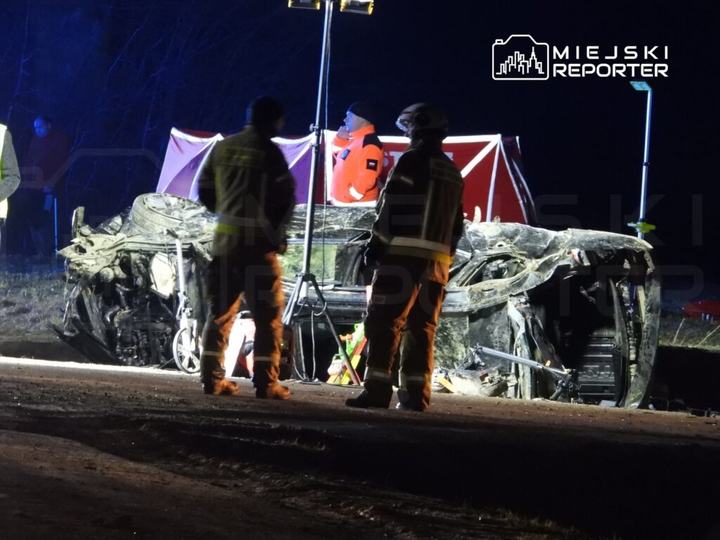 Ratownicy w odblaskowych kamizelkach pracują przy przewróconym samochodzie na drodze w nocy.