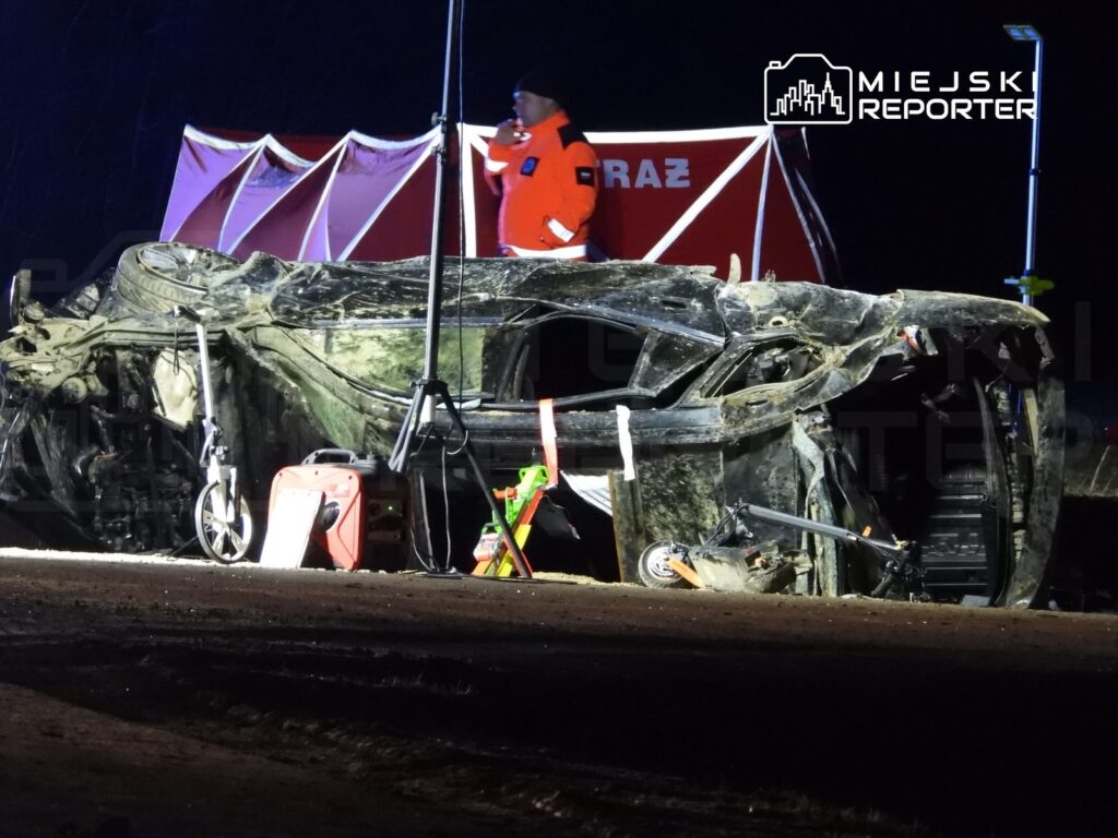 Zniszczony samochód leży na boku na drodze, obok działa ekipa ratunkowa w odblaskowych kamizelkach.