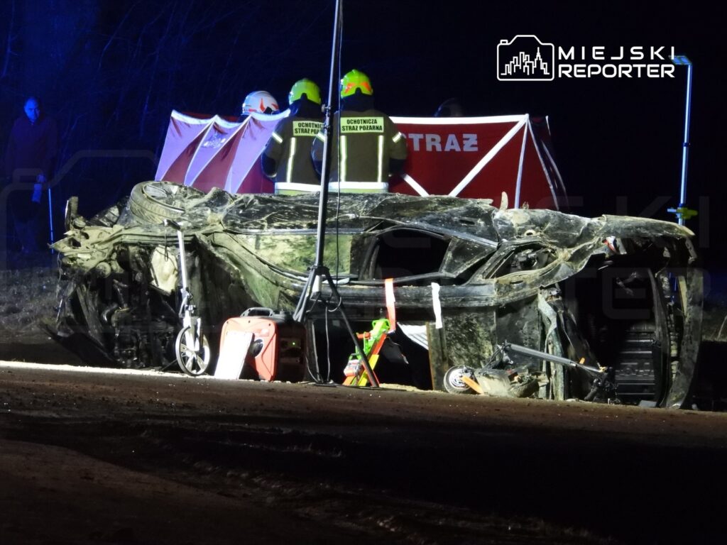 Zniszczony samochód leży na boku na drodze, a w tle widać strażaków pracujących przy oświetleniu awaryjnym.