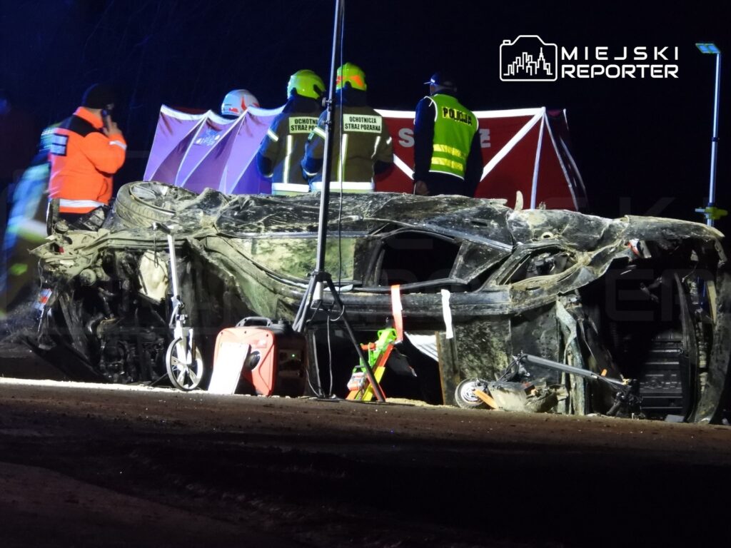Zniszczony samochód leży na boku na drodze, a ratownicy pracują nad zabezpieczeniem miejsca w nocy.