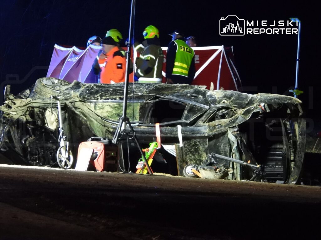 Zniszczony samochód leżący na boku, obok grupy ratowników w odblaskowych kamizelkach, w ciemnej scenerii nocnej.