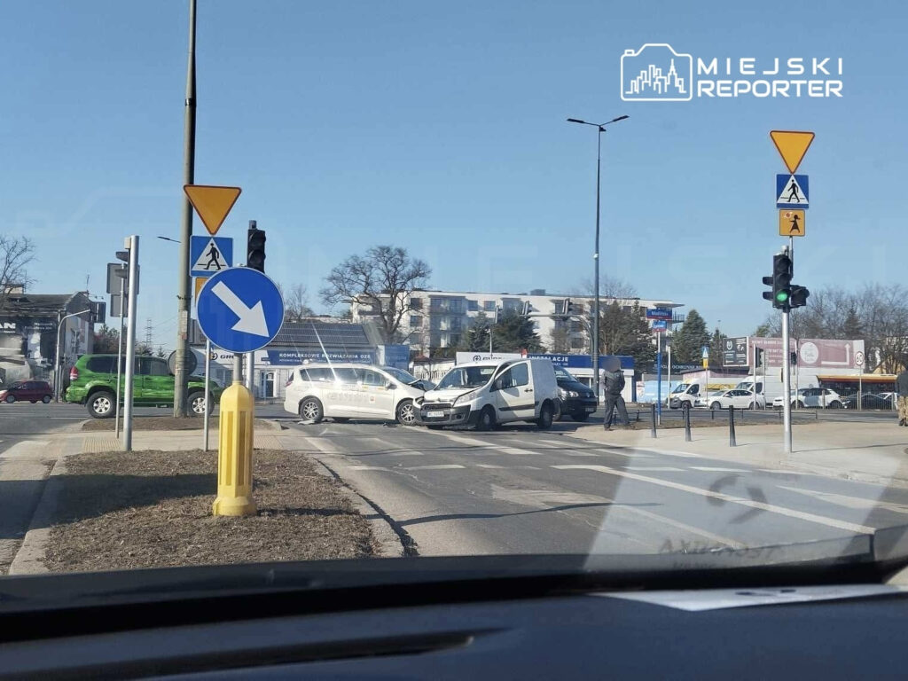 Kierowcy czekają na zielone światło na skrzyżowaniu z ruchem okrężnym, obok znaków drogowych i pojazdów.