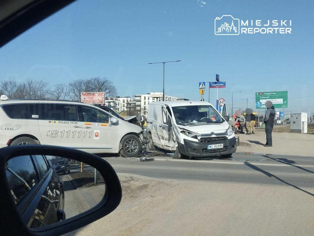 W wyniku kolizji zderzyły się taksówka i dostawczy samochód, na drodze przy znakach drogowych. Obok grupka osób obserwuje sytuację.
