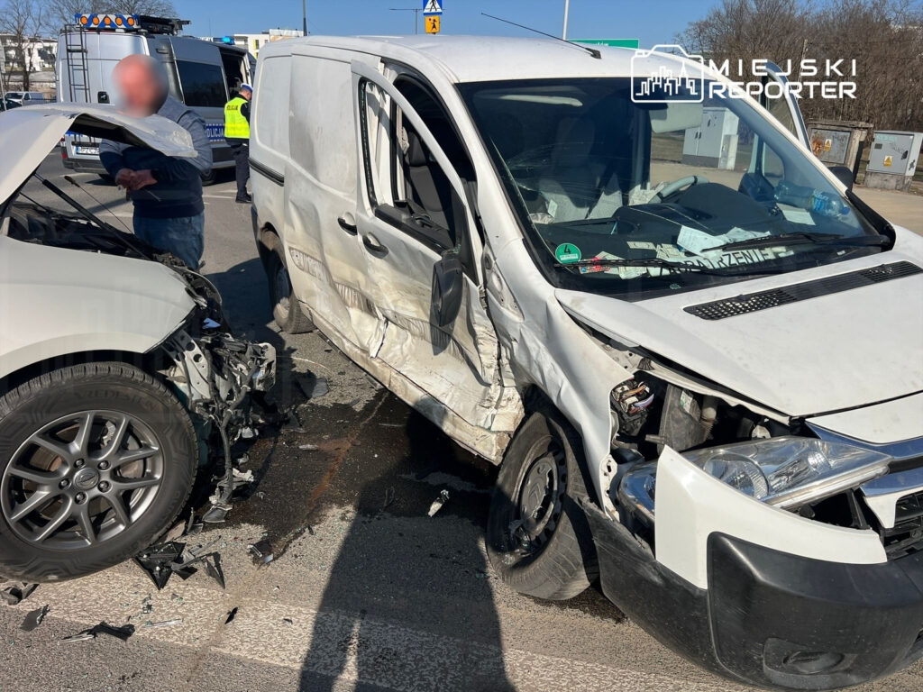 Dwa uszkodzone pojazdy osobowy i dostawczy z widocznymi zniszczeniami na drodze, obok stoi mężczyzna.