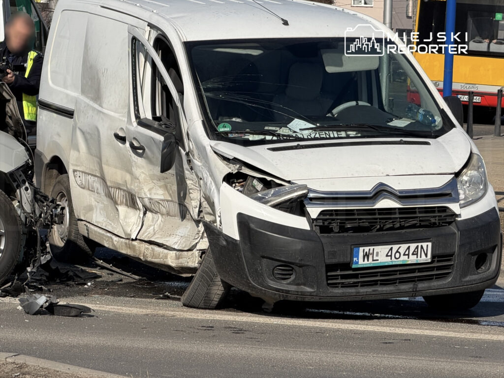 Rozbity biały furgon z uszkodzoną przednią częścią stoi na ulicy, obok policjantów zabezpieczających miejsce zdarzenia.