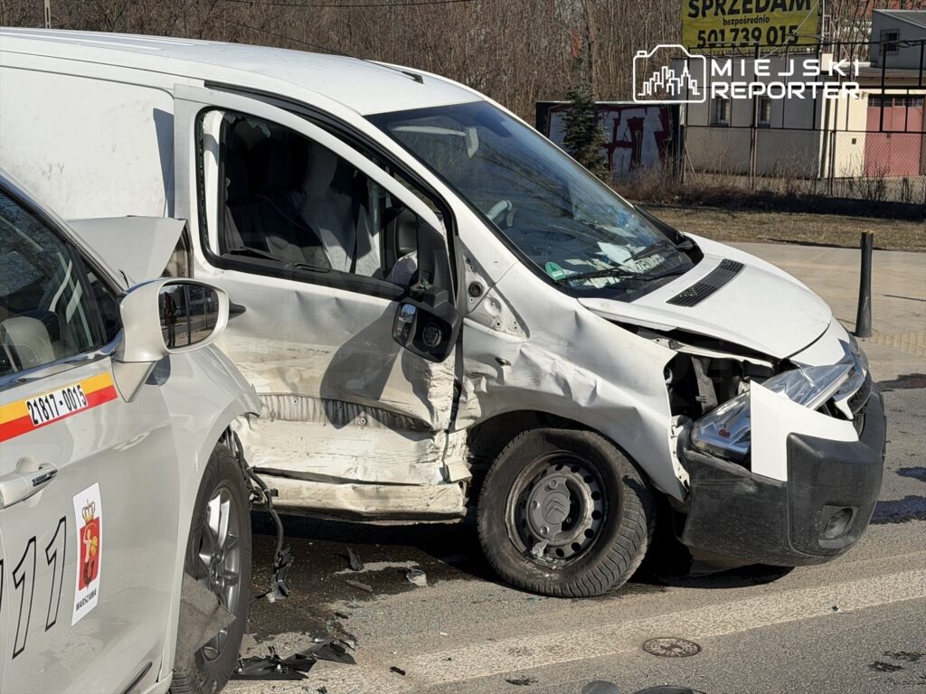Uszkodzony biały furgon z wgniecioną przednią częścią stoi obok radiowozu policyjnego na ulicy.