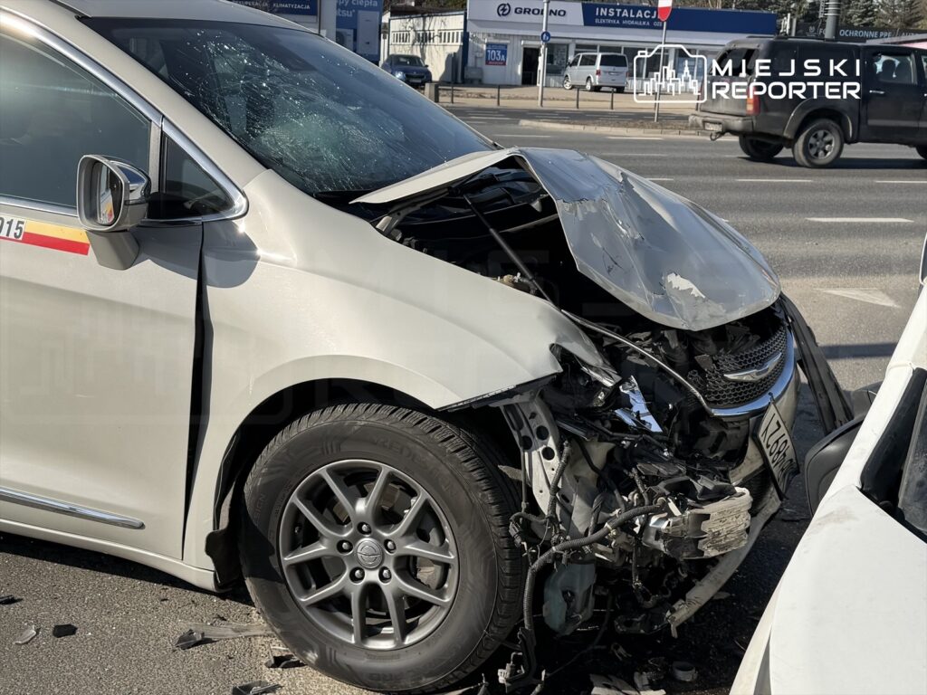 Uszkodzony samochód osobowy z wgniecioną maską i rozbitym przednim szkłem, zaparkowany na ruchliwej ulicy.