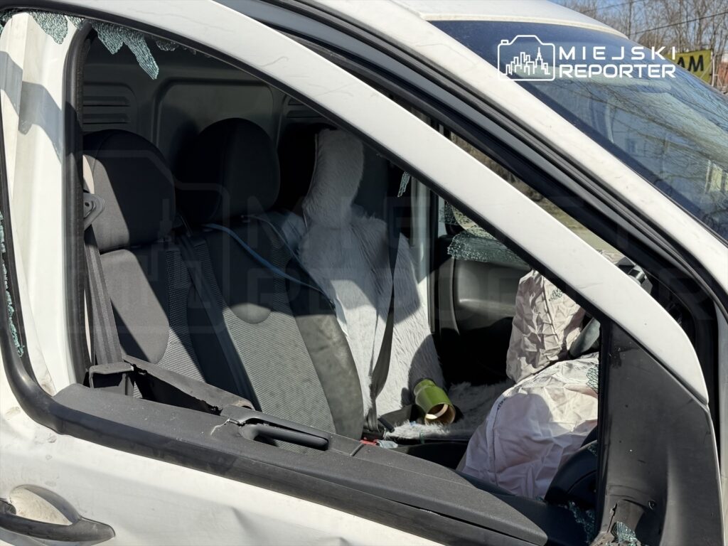 Uszkodzone wnętrze białego samochodu, z wybitymi szybami i uszkodzonymi siedzeniami, stoi w słonecznym otoczeniu.