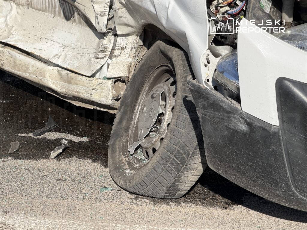 Uszkodzony pojazd z zgniecioną karoserią i uszkodzoną oponą, stojący na drodze. Obok leżą fragmenty blachy.