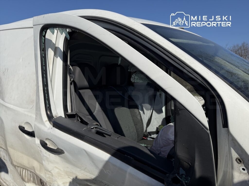 Uszkodzony biały furgon z wybitymi szybami, stojący na drodze w słoneczny dzień. Wnętrze pojazdu widoczne przez otwarte drzwi.