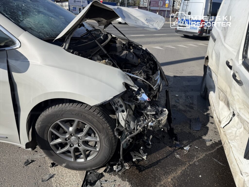 Zniszczony przód osobowego samochodu zaparkowanego na drodze, obok stoi uszkodzony pojazd dostawczy.