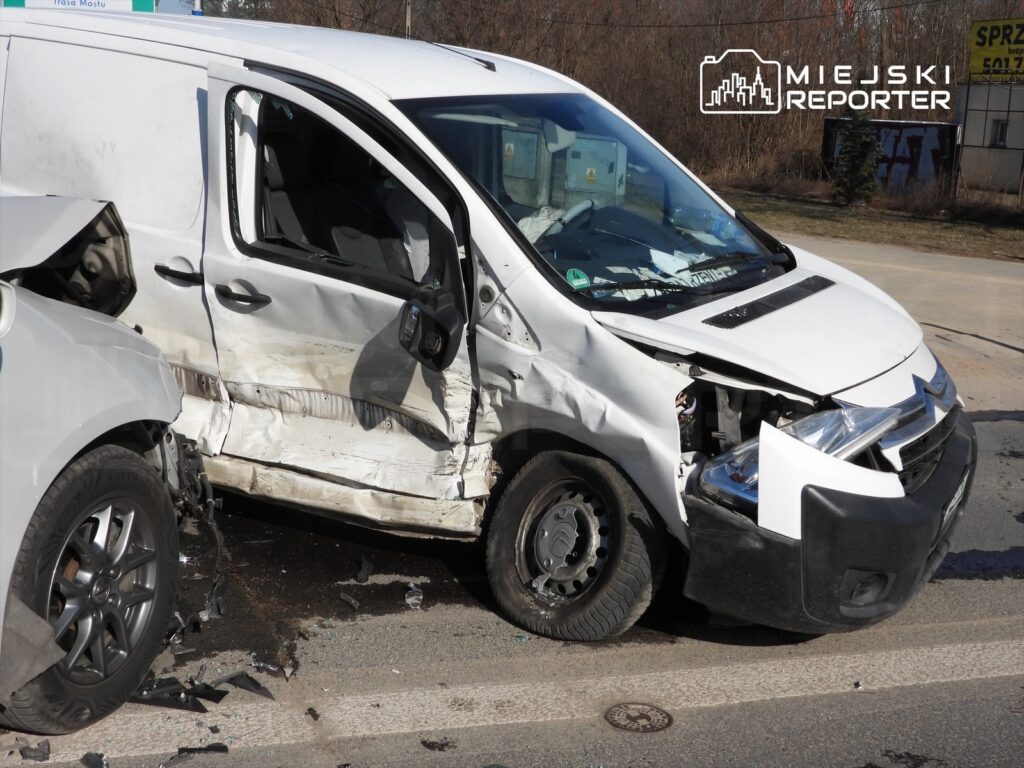 Zniszczony biały furgon z uszkodzoną przednią częścią stoi na drodze, obok widać fragmenty szkła i detali karoserii.