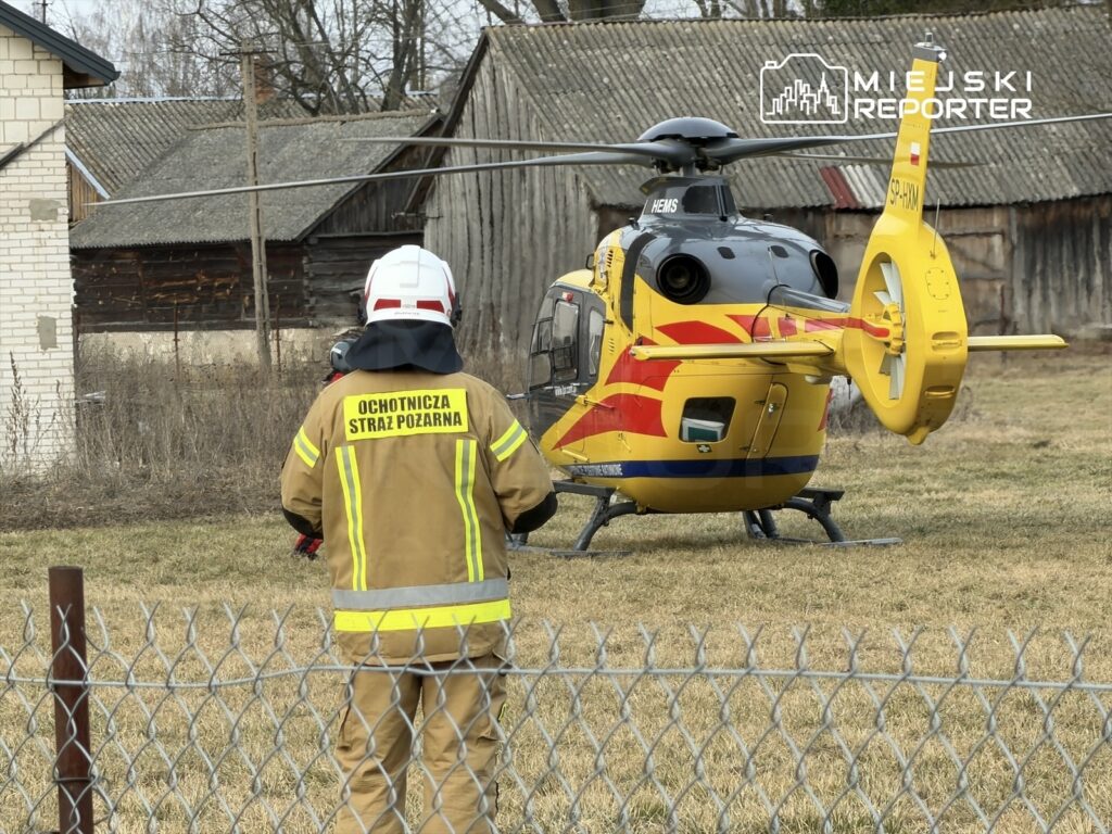 Strażak w mundurze, z plecakiem, obserwuje lądowanie helikoptera medycznego na otwartym terenie.