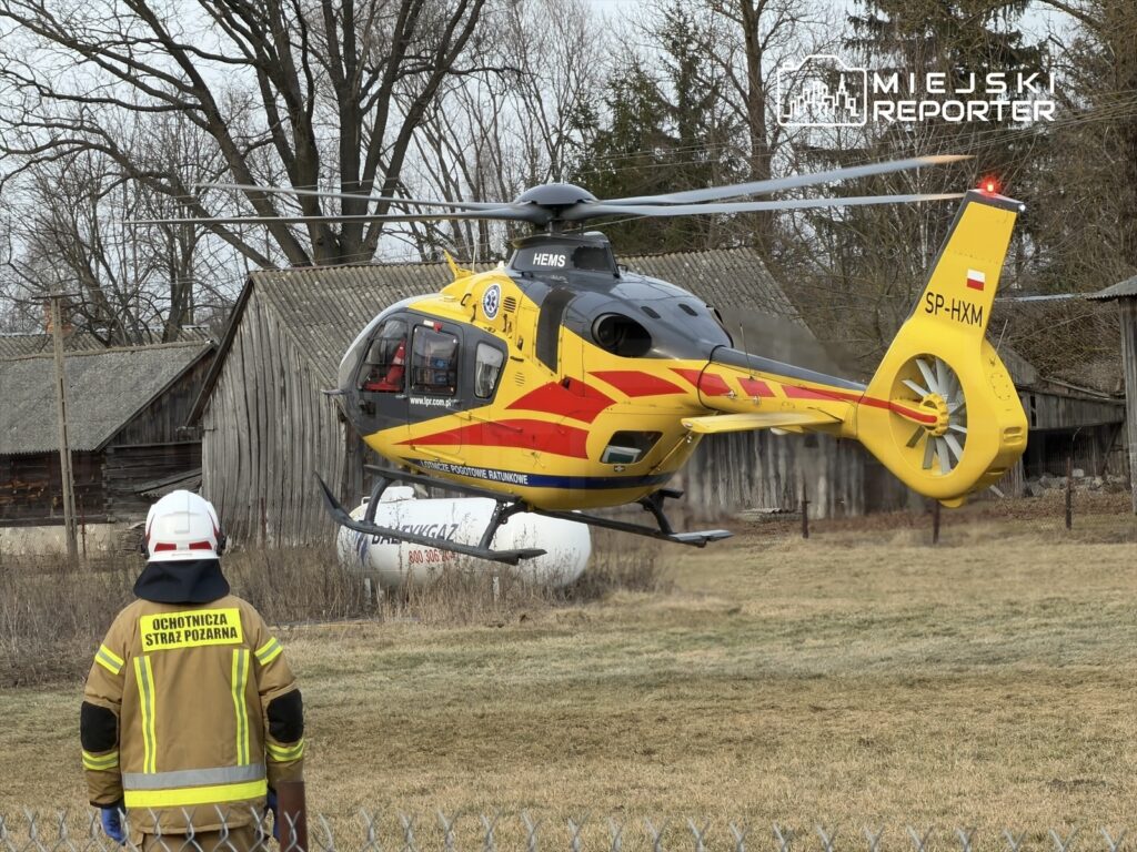 Śmigłowiec ratunkowy ląduje na łące obok budynków gospodarczych, a strażak obserwuje sytuację.