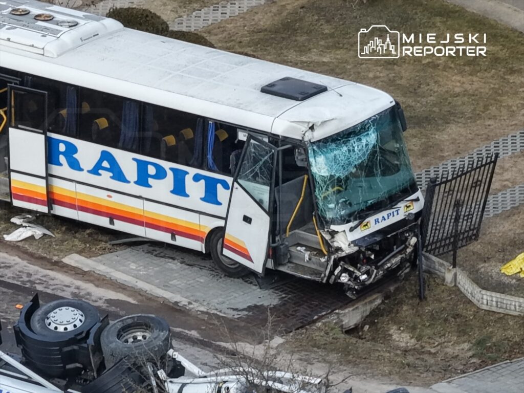 Autobus z widocznymi uszkodzeniami, zatrzymany na chodniku, z rozbitą przednią szybą i uszkodzoną frontową częścią.
