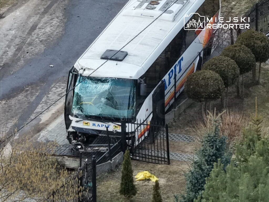 Autobus z rozbitą szybą stoi przy ogrodzeniu, z uszkodzoną przednią częścią, na tle trawnika i drzew.