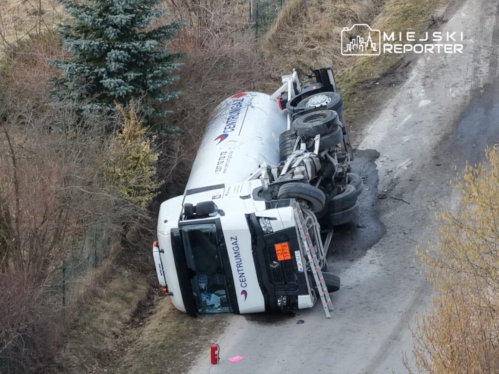 Cysterna z gazem przewrócona na bok na wąskiej drodze w otoczeniu drzew i krzewów.