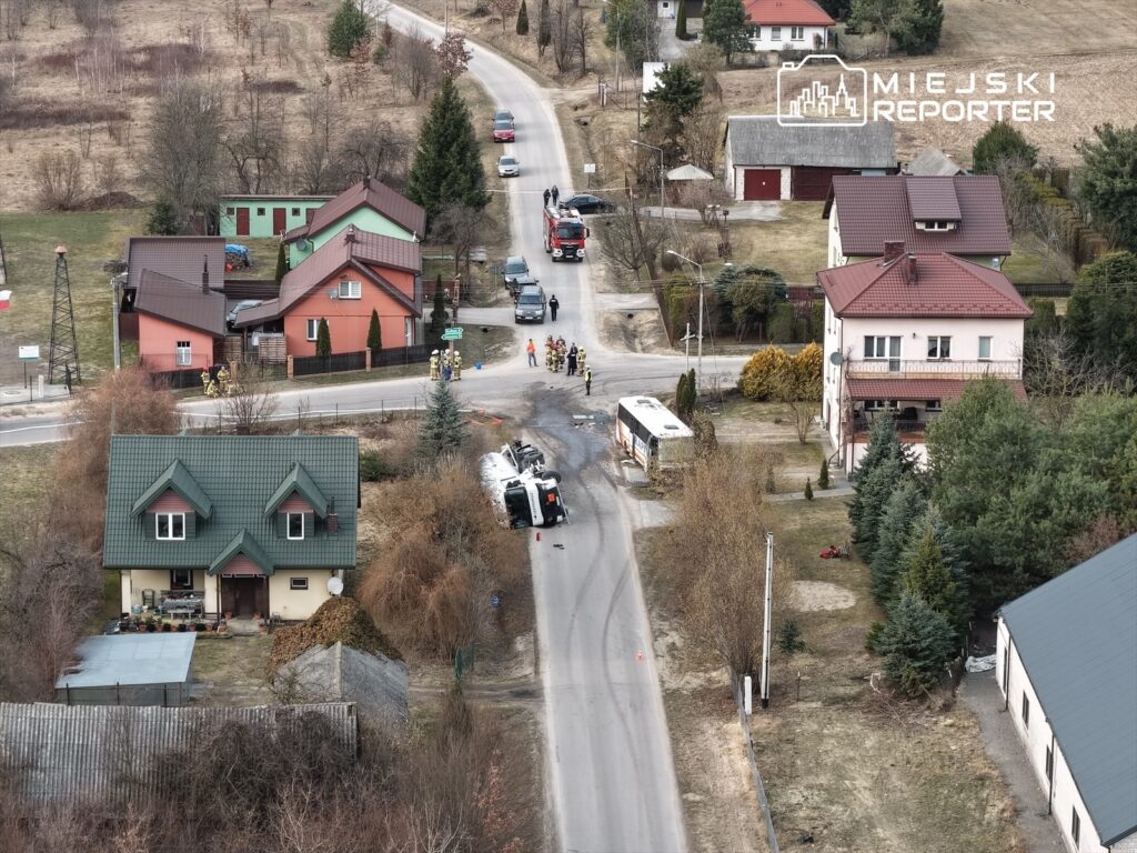 Na drodze leży przewrócony autobus, obok pracują strażacy. W tle widać domy jednorodzinne i zarośla.