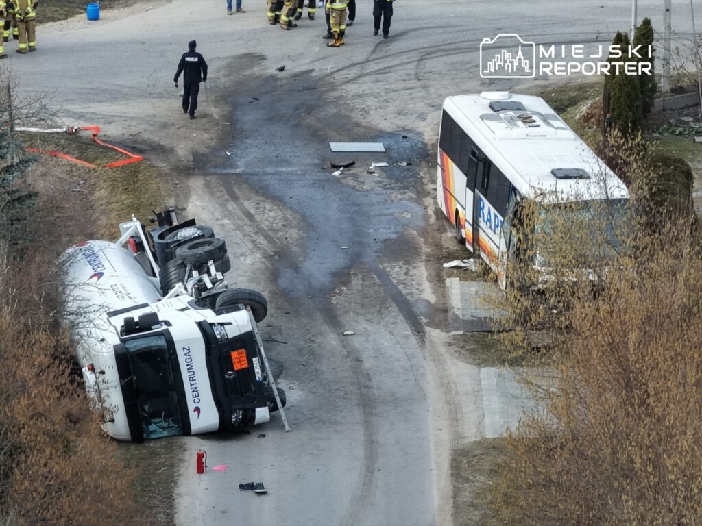 Wielka awaria drogowa z przewróconą cysterną i autobusem, w tle policjanci i strażacy prowadzący akcję ratunkową.