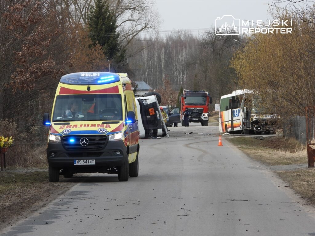Karetka z włączonymi sygnałami świetlnymi zatrzymana na drodze obok przewróconego pojazdu oraz strażaków w akcji.