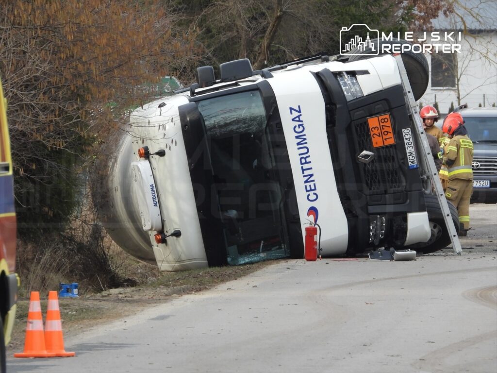 Wielka ciężarówka przewrócona na bok przy drodze, obok której pracują strażacy w mundurach.