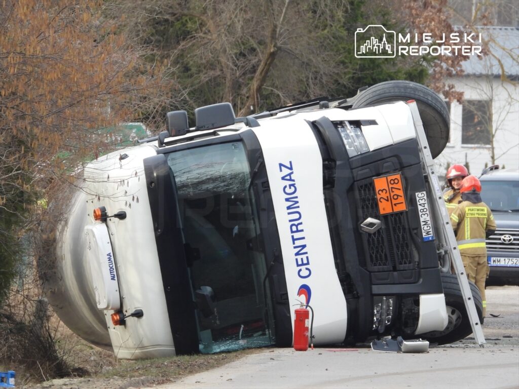 Cysterna przewożąca gaz leży na boku na poboczu drogi, obok interweniują strażacy w sprzęcie ochronnym.
