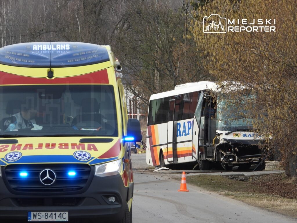 Ambulans z włączonymi sygnałami świetlnymi stoi na drodze obok uszkodzonego autobusu z widocznym napisem "RAP".
