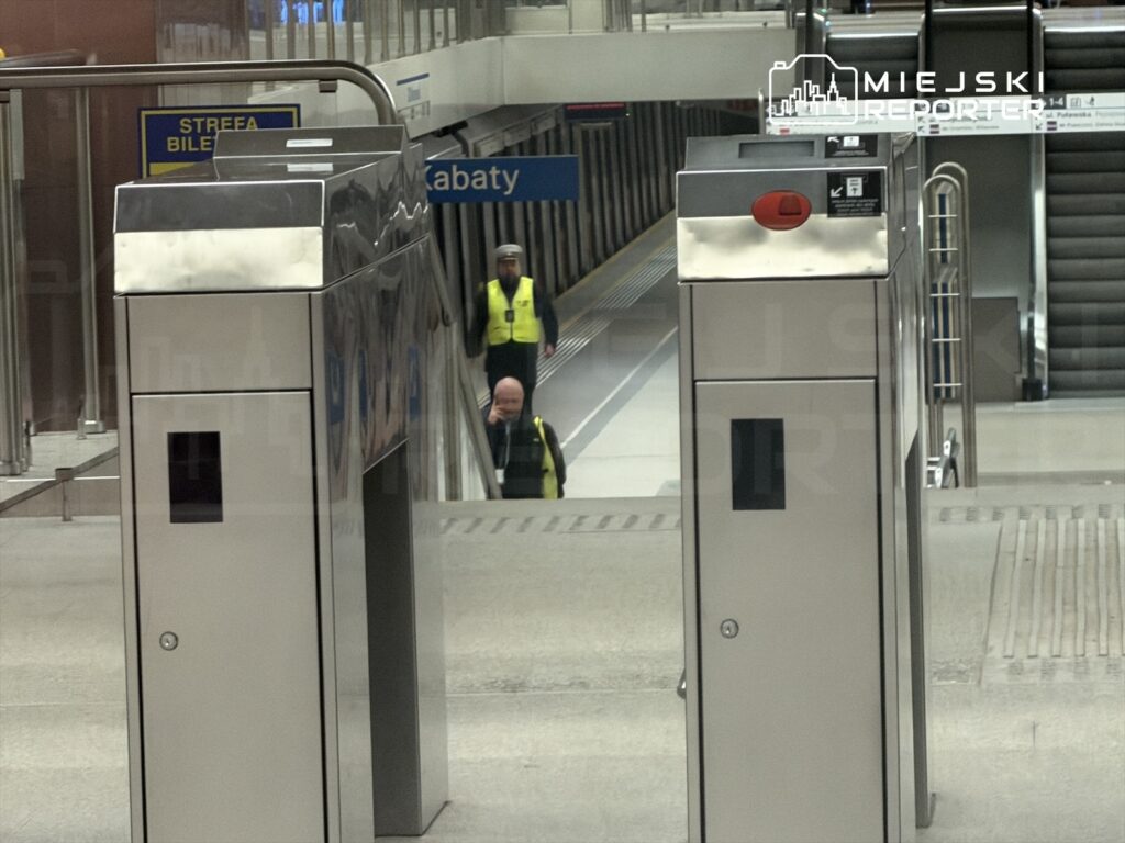 Mężczyzna w żółtej kamizelce przechodzi obok bramek biletowych na stacji metra. W tle widoczne perony.