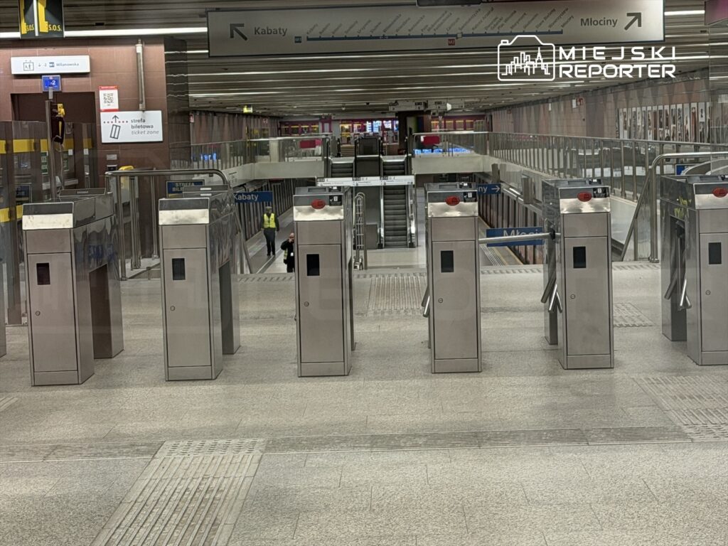 Pojedyncze bramki biletowe w nowoczesnej stacji metra, w tle widoczny personel w żółtej kamizelce.