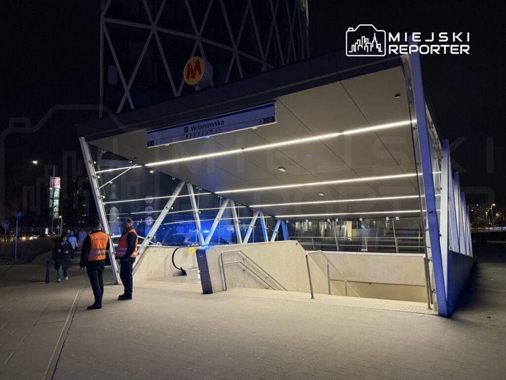 Mężczyźni w odblaskowych kamizelkach stoją przed nowoczesnym wejściem do stacji metra w nocy.