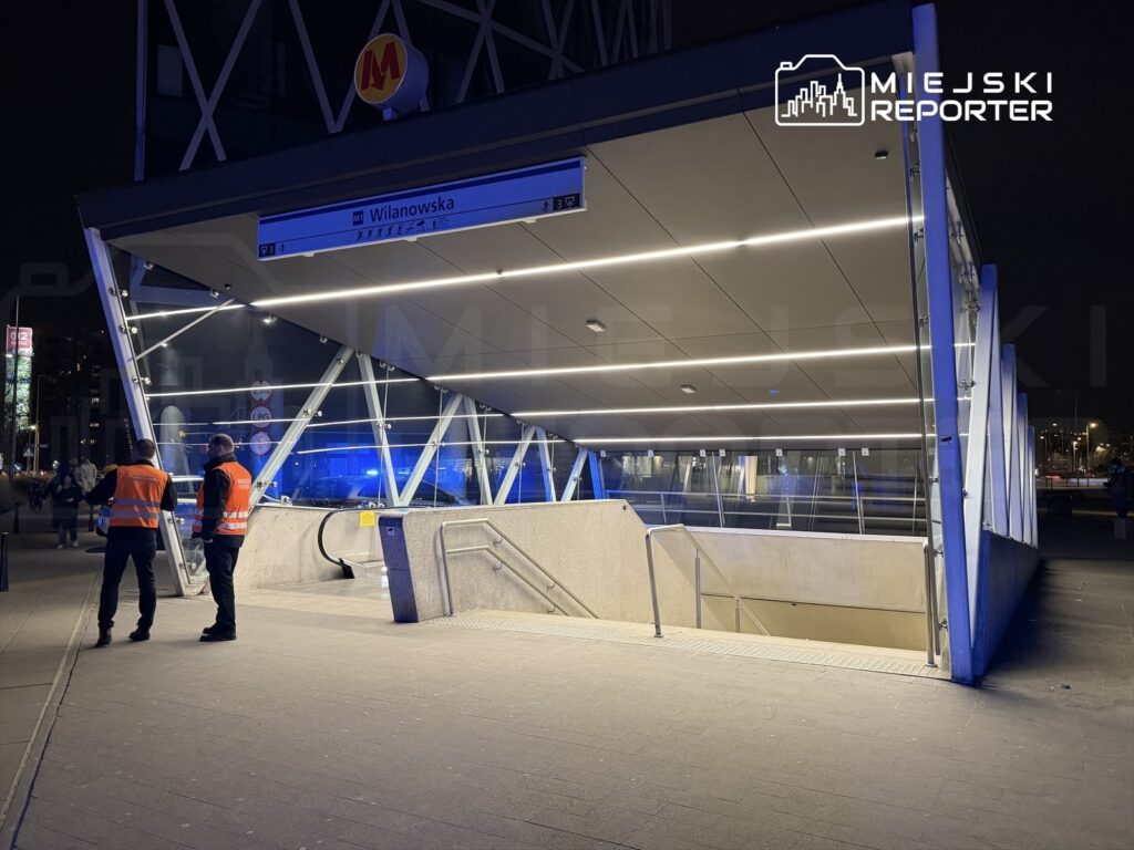 Dwaj mężczyźni w pomarańczowych kamizelkach rozmawiają przed wejściem do stacji metra w nocy.