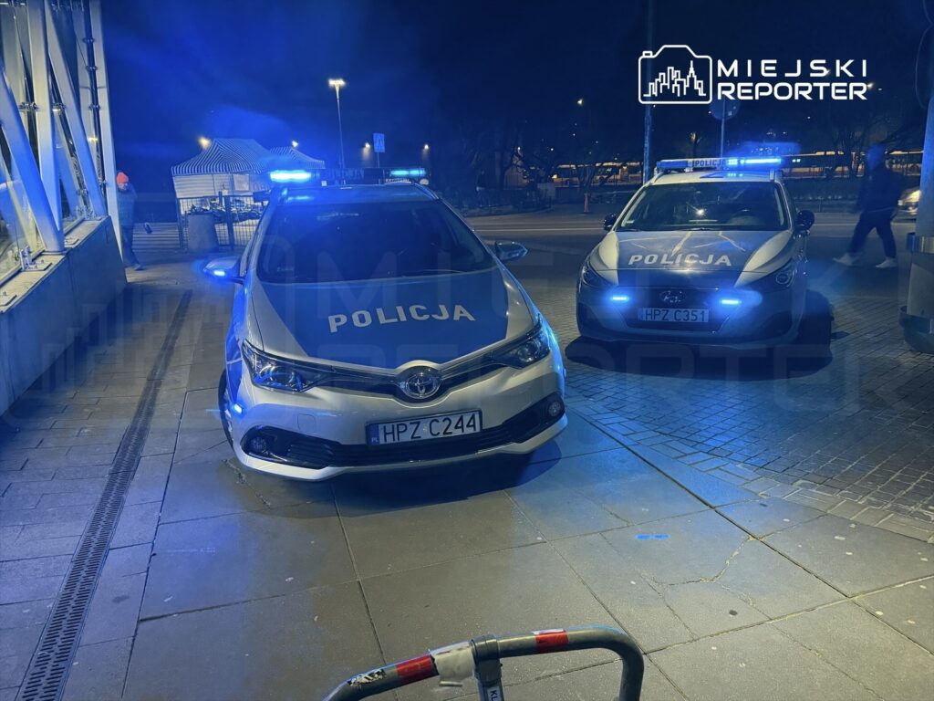 Dwa radiowozy policyjne zaparkowane na oświetlonym chodniku, emitujące niebieskie światła.