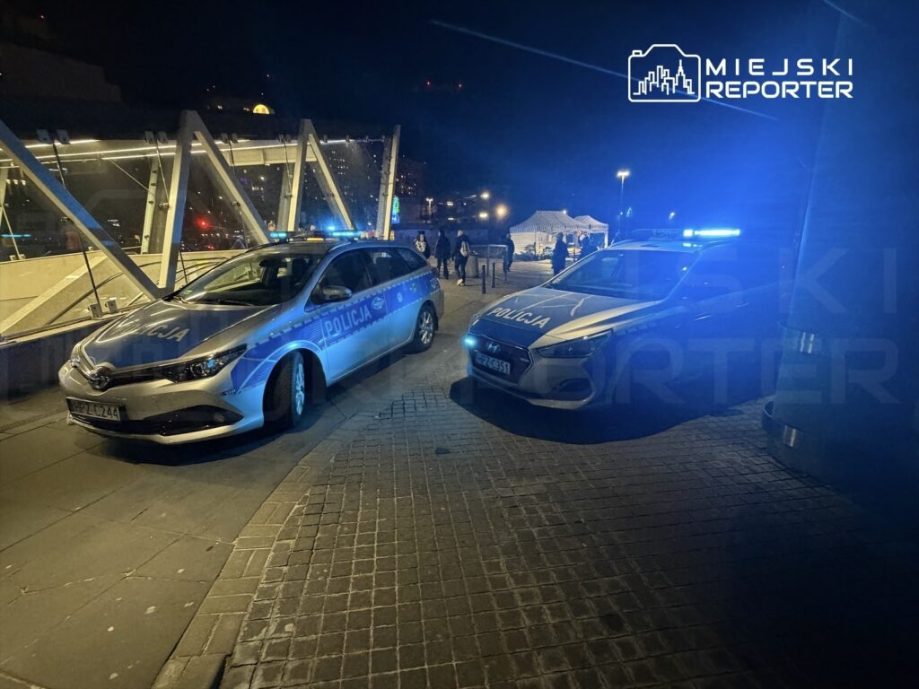 Dwa radiowozy policyjne z włączonymi sygnałami świetlnymi stoją na chodniku w nocy, w tle widoczna architektura.