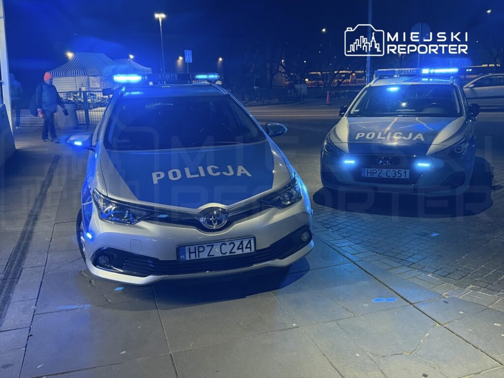 Dwa radiowozy policyjne z włączonymi sygnałami świetlnymi stoją na oświetlonym placu nocą, obok przechodnia.