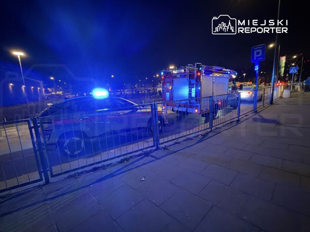 Policyjny radiowóz i wóz strażacki z włączonymi sygnałami świetlnymi zaparkowane na miejskiej ulicy w nocy.