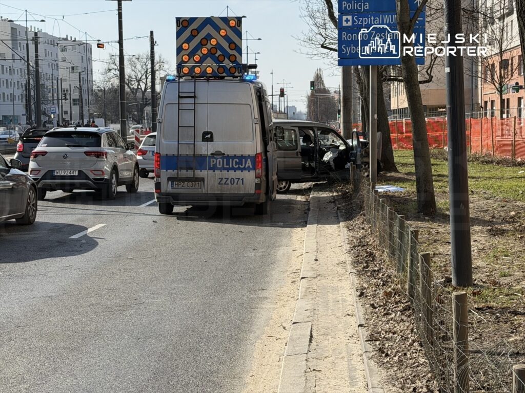 Policyjny radiowóz z włączonymi sygnałami świetlnymi zatrzymany na ulicy obok uszkodzonego samochodu osobowego.