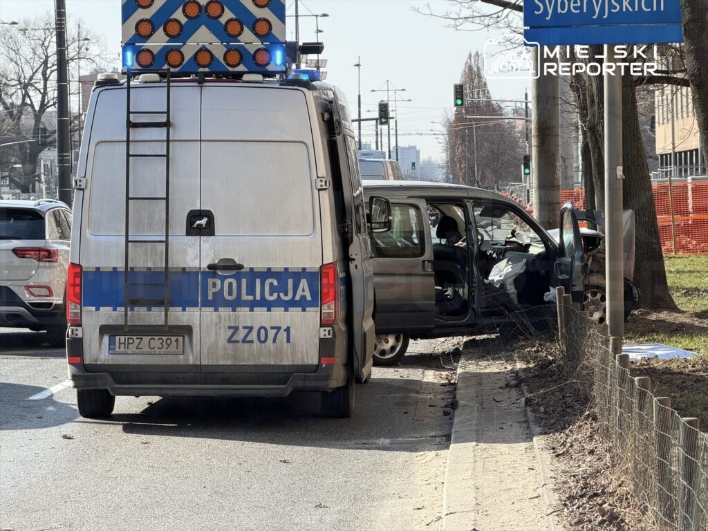 Policyjny van z włączonymi sygnałami świetlnymi stoi obok uszkodzonego pojazdu na ulicy, w tle widoczne sygnalizatory.