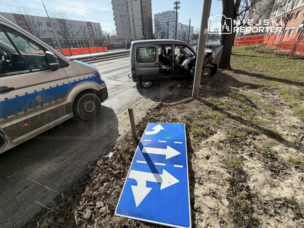 Uszkodzony samochód osobowy stoi na poboczu, obok policyjny furgon, a na ziemi leży nieczytelny znak drogowy.