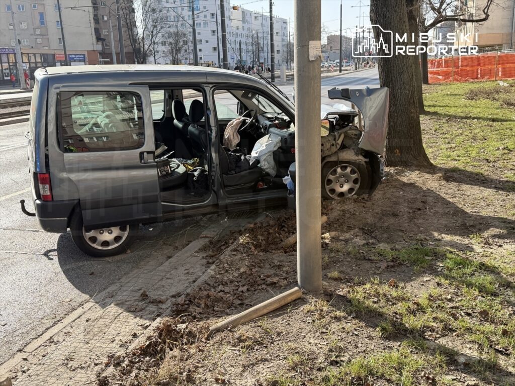 Uszkodzony samochód dostawczy stoi przy latarni, z otwartymi drzwiami i widocznymi uszkodzeniami przodu.