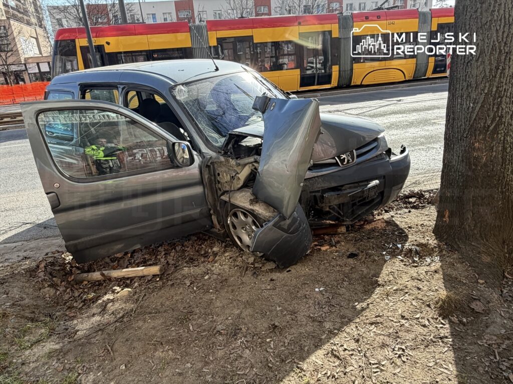 Uszkodzony samochód osobowy stoi na poboczu, z otwartymi drzwiami, obok drzewa, w tle widoczny tramwaj.
