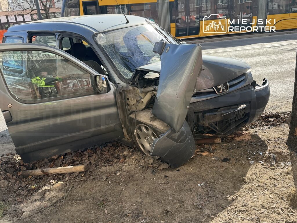 Uszkodzony samochód osobowy stoi na poboczu drogi, z otwartymi drzwiami i widocznymi uszkodzeniami przodu.
