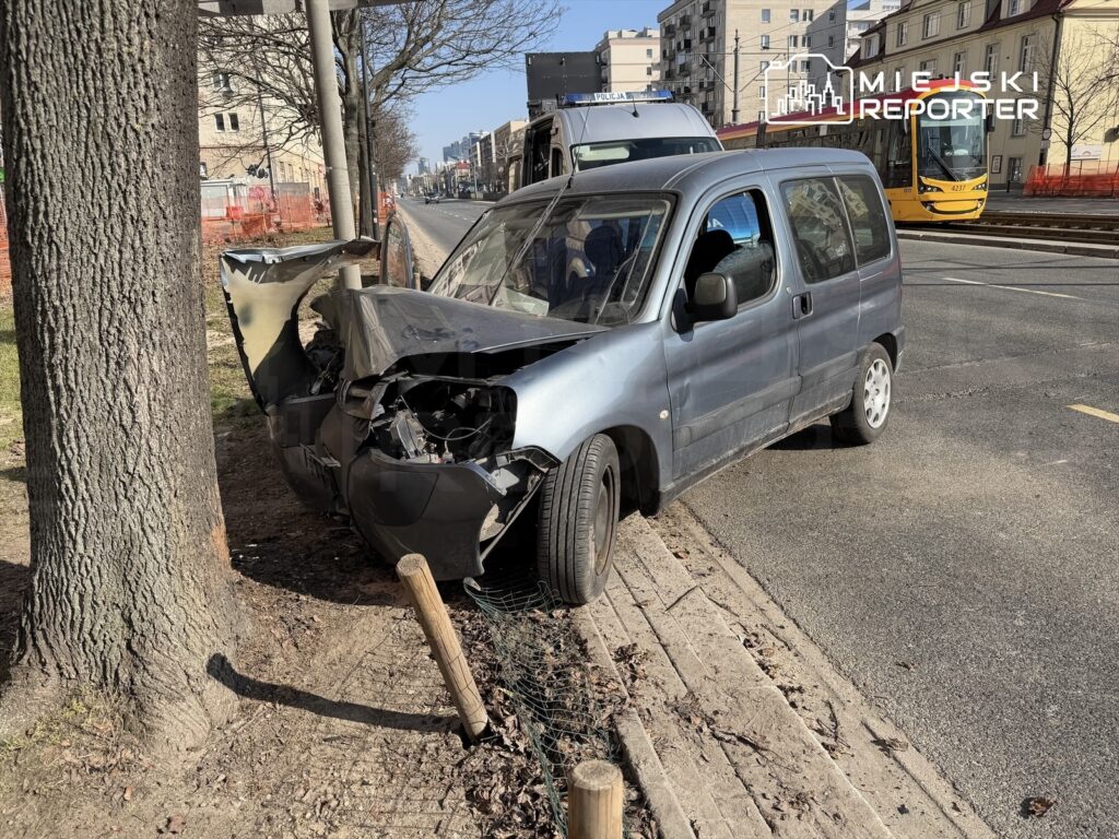 Uszkodzony samochód dostawczy stoi przy drodze, zderzony z drzewem, obok torów tramwajowych w miejskim otoczeniu.