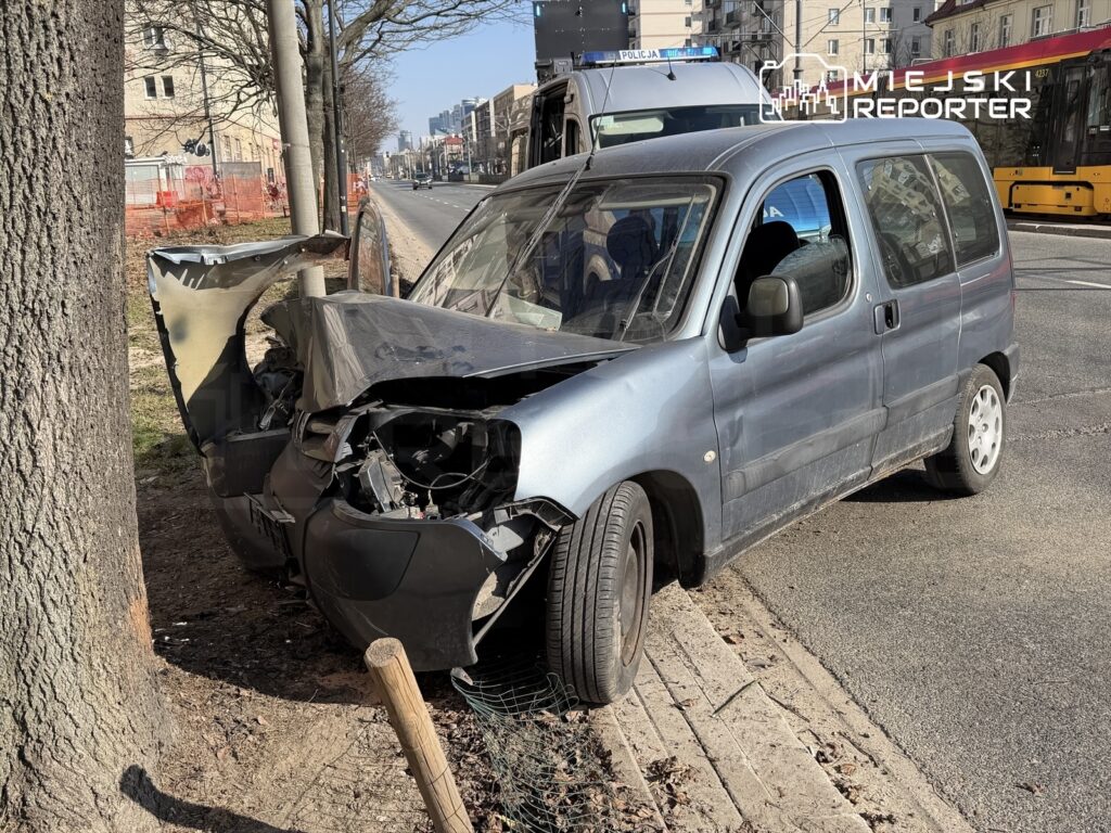 Uszkodzony samochód osobowy z wgniecioną przednią częścią stoi przy chodniku, obok drzewa, na ruchliwej ulicy.