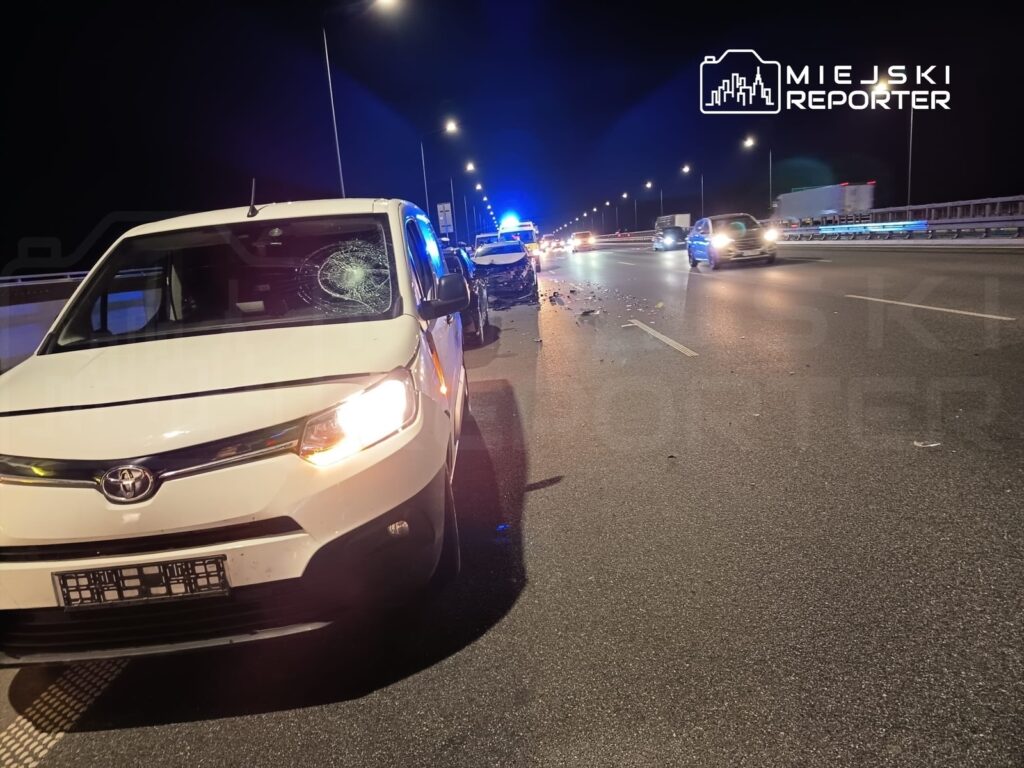 Biały samochód osobowy z uszkodzoną szybą stoi na drodze, obok widać policyjne światła i ruch innych pojazdów.