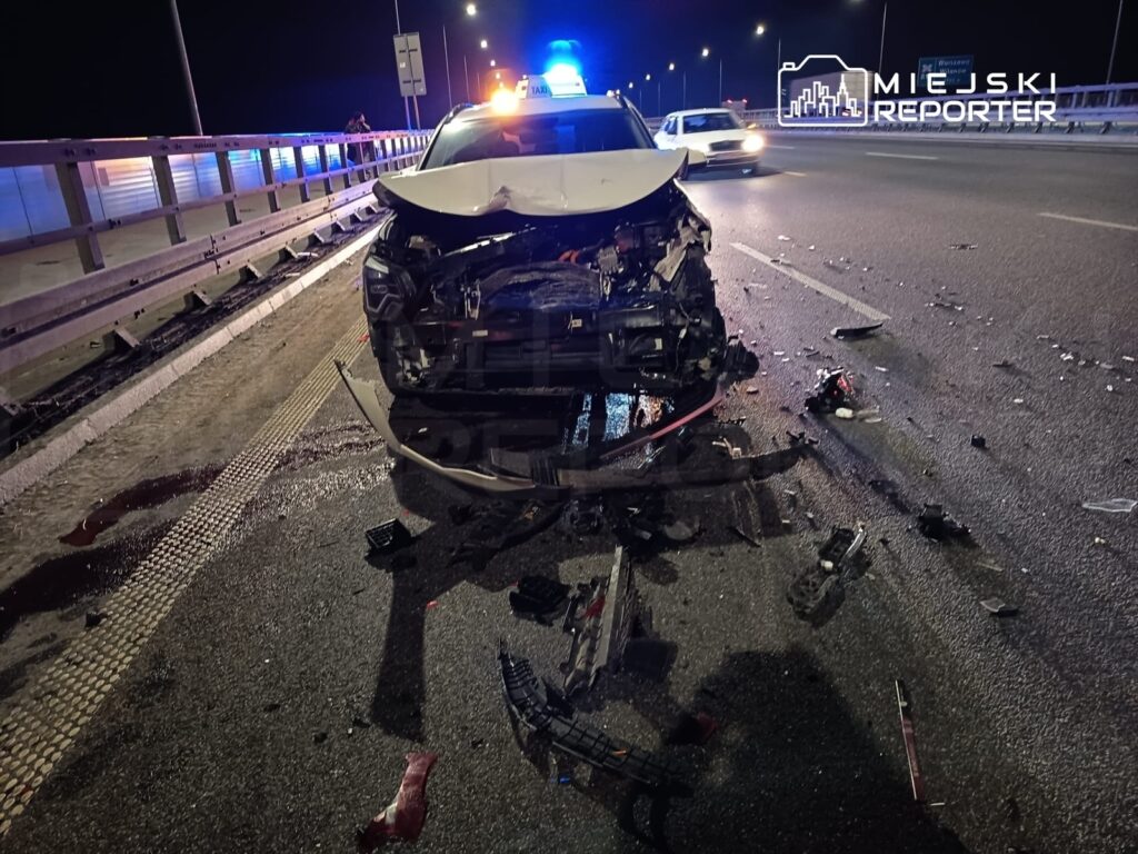 Uszkodzony radiowóz policyjny z rozbitym przodem, otoczony fragmentami karoserii na oświetlonej drodze nocą.