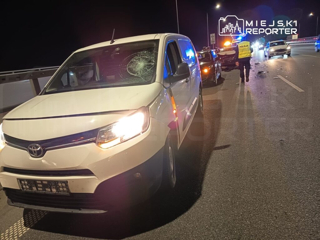Biały samochód osobowy z rozbityą szybą stoi na drodze, obok widoczne pojazdy policyjne oraz funkcjonariusz w kamizelce odblaskowej.