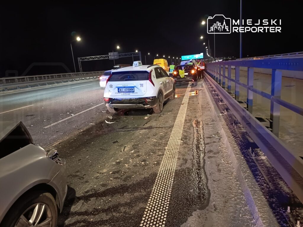 W nocy na drodze widać uszkodzone samochody po kolizji, obok pracują służby ratunkowe.