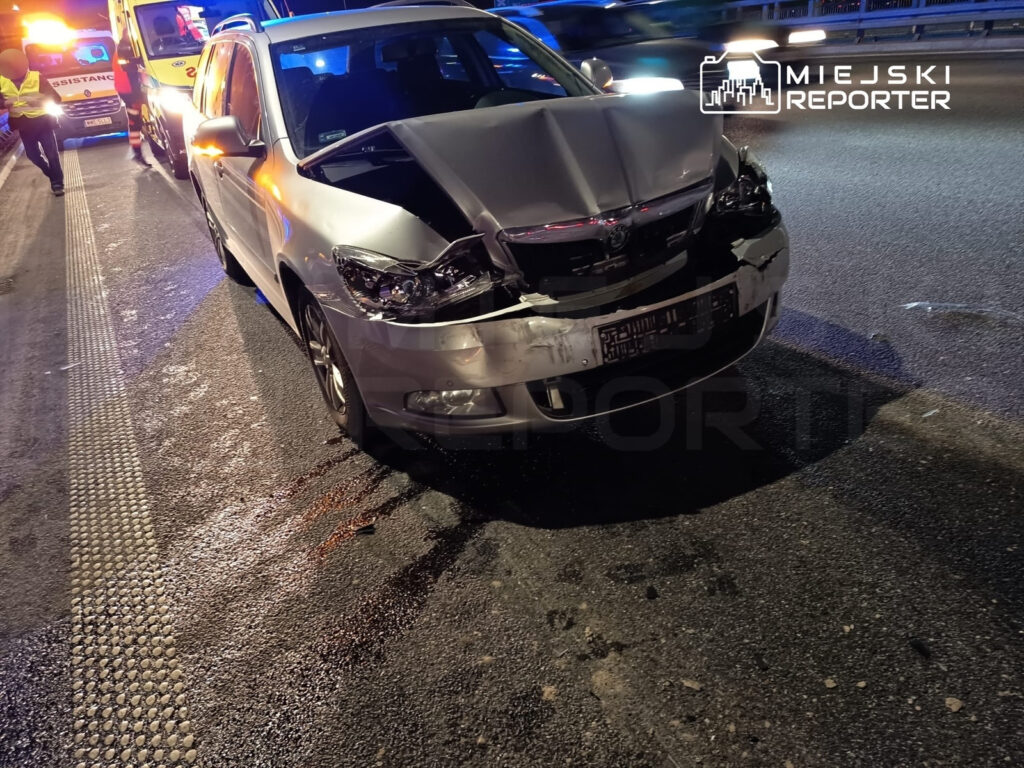 Uszkodzony samochód osobowy z wgniecionym przodem stoi na drodze, obok widać pojazdy służb ratunkowych.