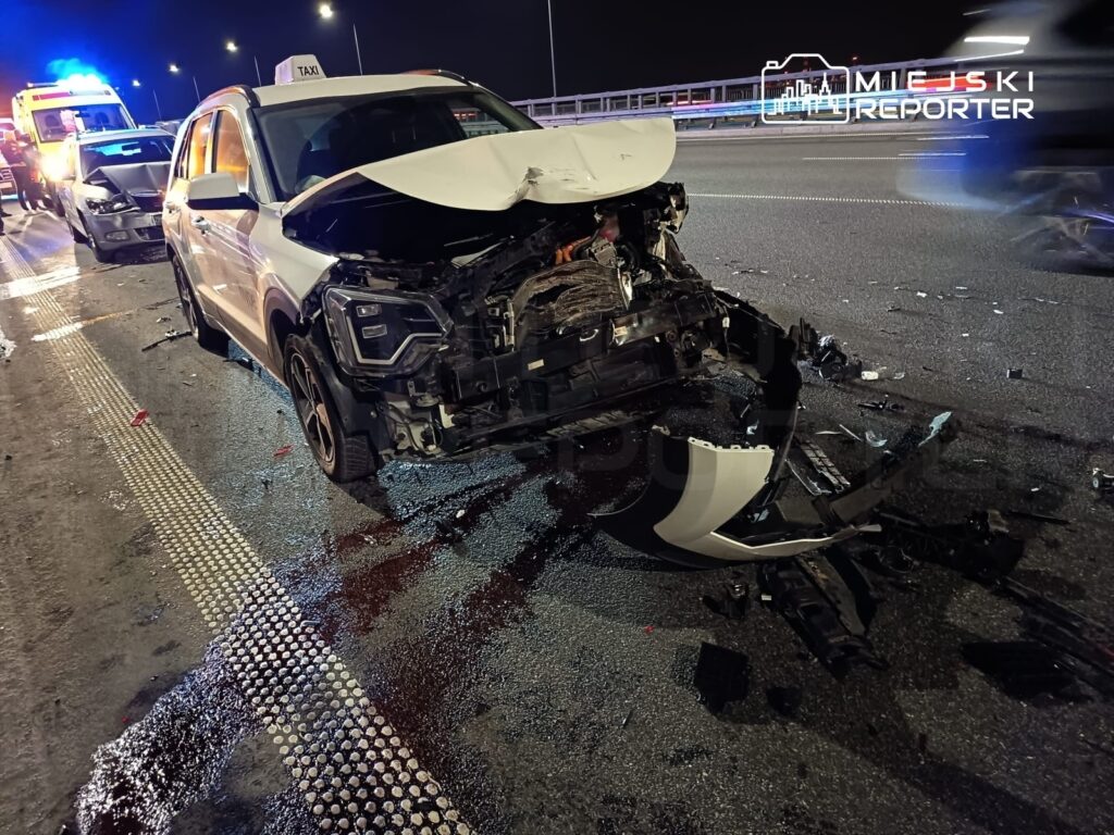 Zniszczony samochód osobowy na drodze, obok niego widać pozostałości wypadku oraz włączone światła sygnalizacyjne pojazdów ratunkowych.