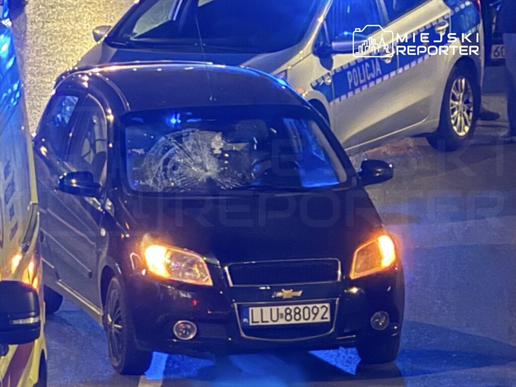 Czarny samochód osobowy z wybitym przednim szybą stoi na ulicy, obok pojazdów policyjnych z włączonymi sygnałami świetlnymi.